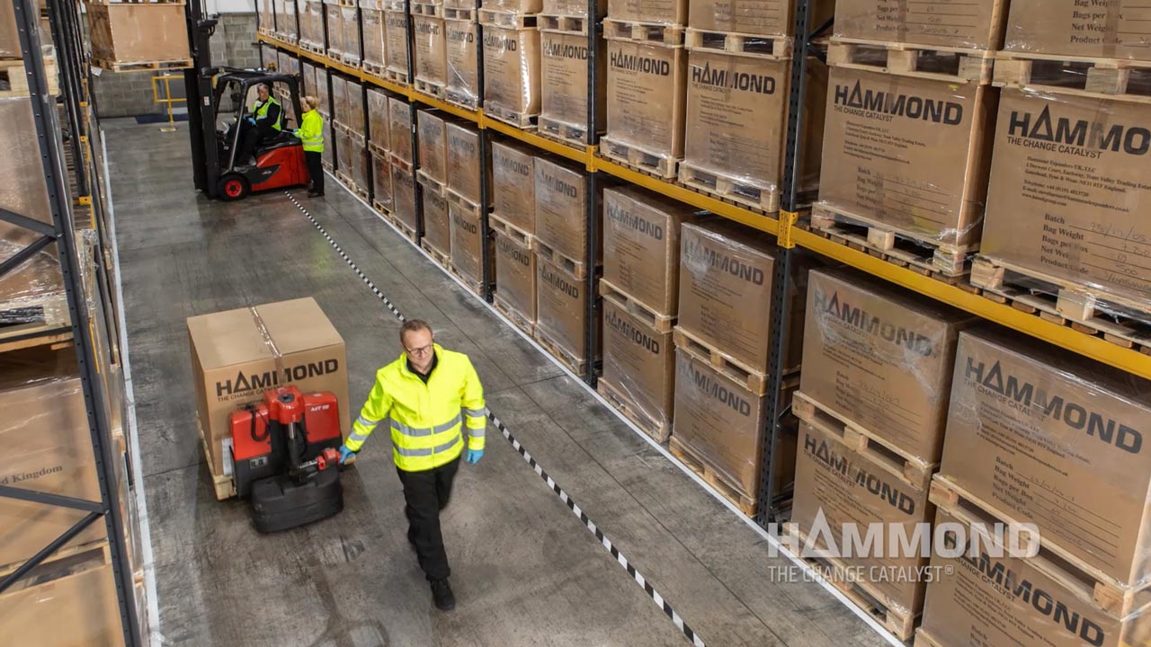 Man pulling large box on pallet in isle of many boxes stacked to the ceiling of warehouse.